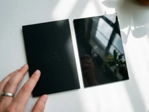 Two business cards on white surface showing matte black and glossy finishes, designer's hand touching matte card corner