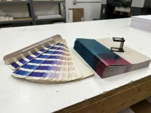 Pantone kleurenwaaier naast gedrukte brochures op een drukkerstafel, met een loupe en levendige CMYK-inkttinten in studioverlichting.