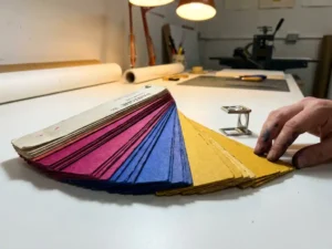 Pantone kleurenwaaier uitgespreid op wit werktafel met loupe, inktgevlekte vingers op kleurchip, warme studiobelichting.